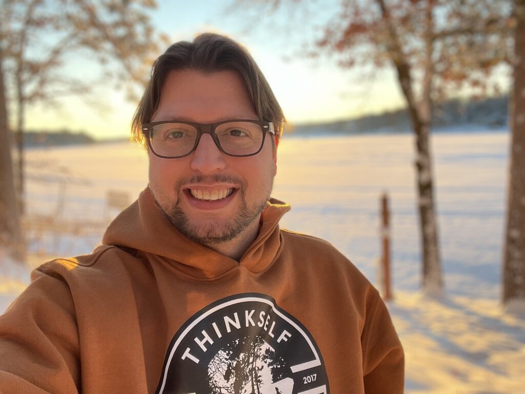 A person in a "ThinkSelf" hoodie smiles at the camera with a snowy landscape and trees in the background during sunset.