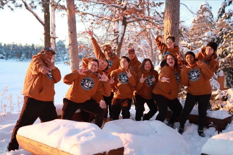 A group of people wearing matching "ThinkSelf" hoodies poses energetically in a snowy outdoor setting with trees and a sunset in the background.