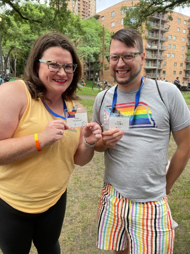 The image features two individuals smiling and displaying their name badges at an outdoor event. The woman on the left has medium-length dark hair and wears glasses, a yellow sleeveless top, and black leggings. The man on the right sports glasses, a gray T-shirt, and multicolored striped shorts. The background shows a park setting with trees and an apartment building, suggesting a community or social gathering. They both wear lanyards indicating affiliation with "ThinkSelf".