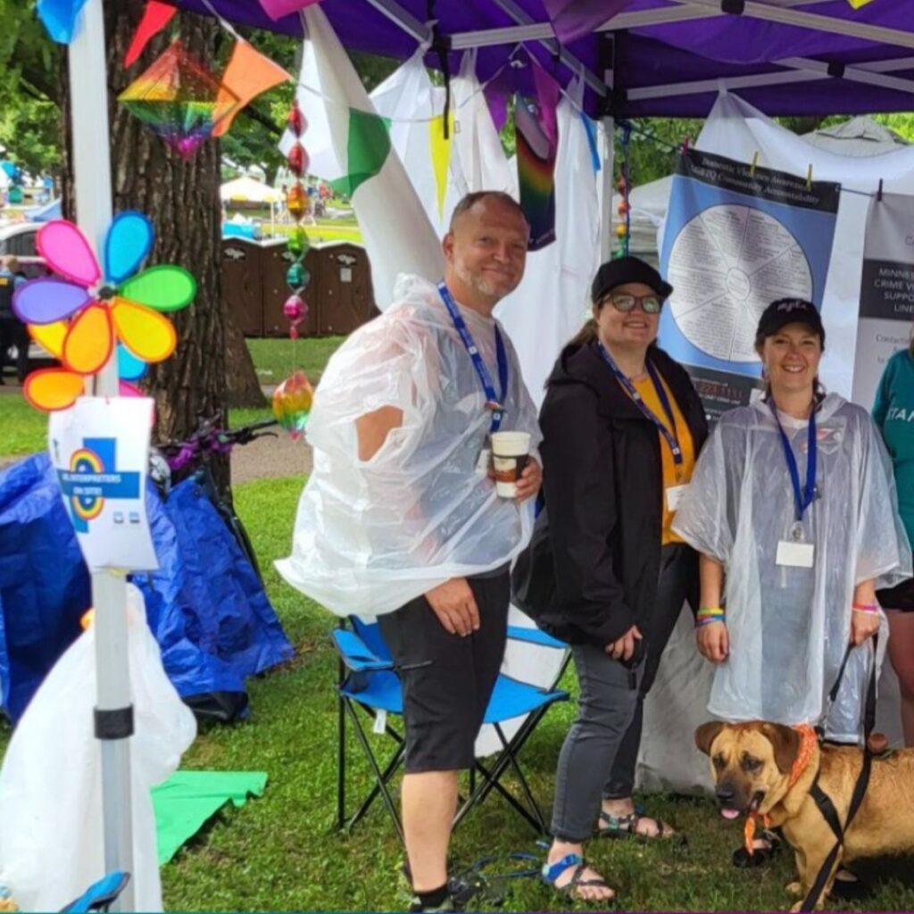 The image shows three adults and a dog under a colorful canopy tent adorned with rainbow flags and banners. The tent displays various informational posters and handouts. Two of the individuals are wearing clear rain ponchos, and the third wears a dark jacket.