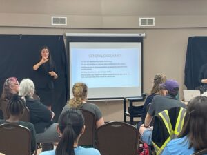 The image shows a woman presenting in front of a room filled with attendees. She is standing beside a screen displaying a slide titled "GENERAL DISCLAIMER." The presenter, using American Sign Language (ASL), appears to be communicating the content of the slide to the audience. The audience, diverse in age and appearance, is seated facing the presenter, suggesting an engaged and attentive atmosphere at this educational or informational event.