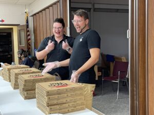 The image depicts two people, a man and a woman, behind a counter, both wearing black shirts with the ThinkSelf logo. They are smiling and giving a thumbs-up gesture while wearing gloves, standing behind stacks of pizza boxes.