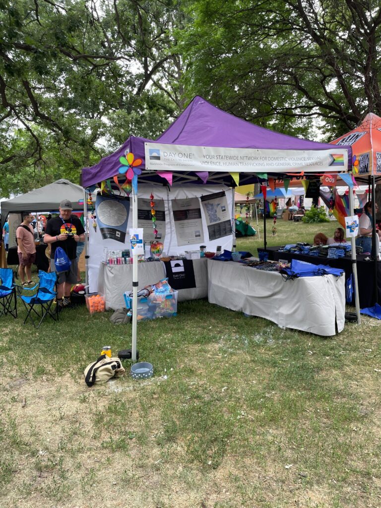 The image depicts an outdoor event booth under a purple canopy, surrounded by trees and other festival stands. The booth is adorned with colorful banners, informational posters, and various handouts on a table.