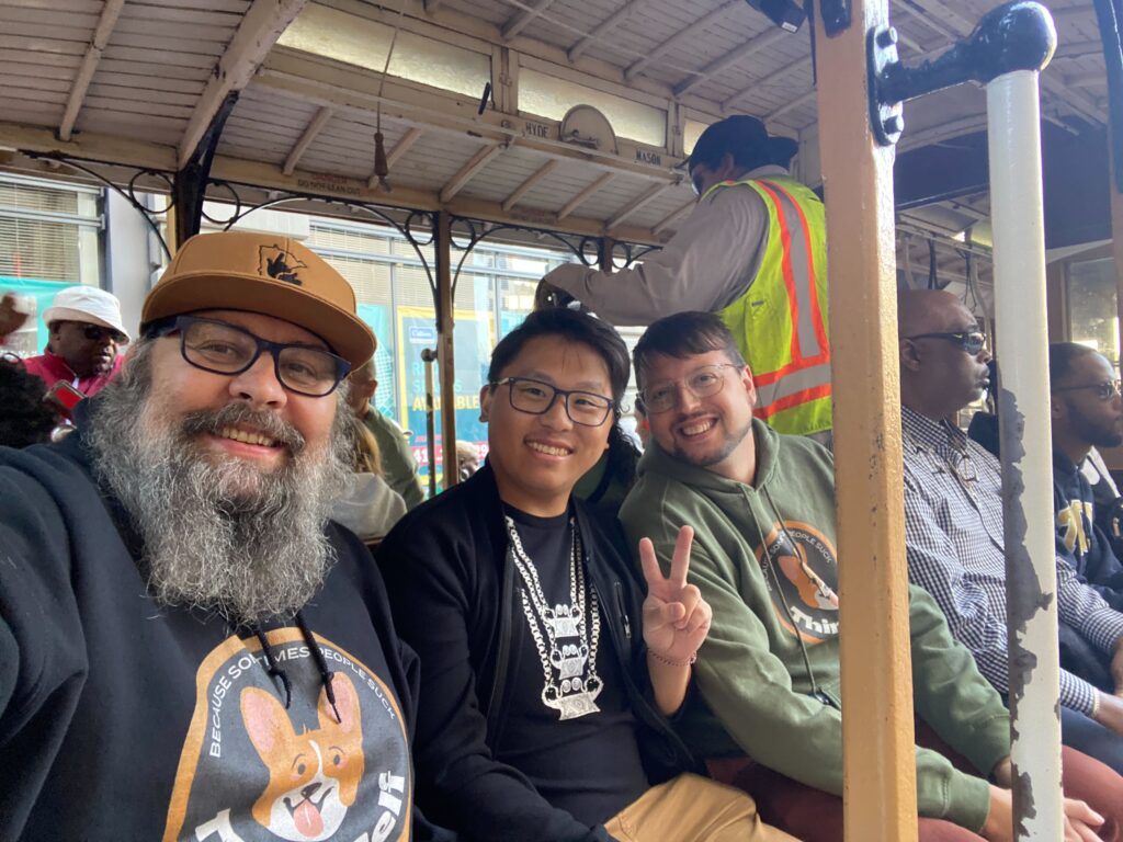 The image depicts three individuals riding on a cable car. The person on the left, a man with a grey beard and glasses, is wearing a tan hat and a black hoodie featuring a rabbit design, and is smiling at the camera. The person in the middle, wearing glasses and a black hoodie, is making a peace sign with their hand. The person on the right, in a green jacket and glasses, is also smiling and making a peace sign. The background shows other passengers and the cable car operator.