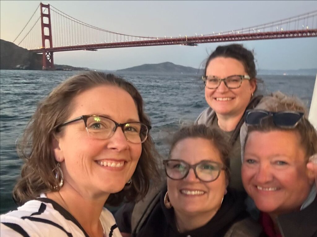 The image shows a group of four women posing for a selfie with the Golden Gate Bridge in the background. They appear to be on a boat or near the water, given the perspective and the way the water and bridge are visible behind them. The women are smiling and seem to be enjoying their outing, capturing the moment in a casual, joyful manner.