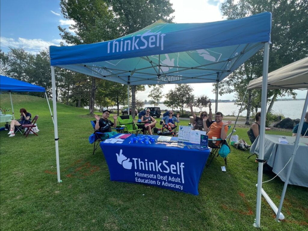 The image shows an outdoor event under a large blue canopy with the "ThinkSelf" logo. There are several people seated around, some interacting with each other, others lounging and enjoying the day. The setting is a grassy park by a lake, providing a relaxed and scenic backdrop. The booth is equipped with informational materials and promotional items on a table, indicating it is an outreach or community engagement effort by ThinkSelf.