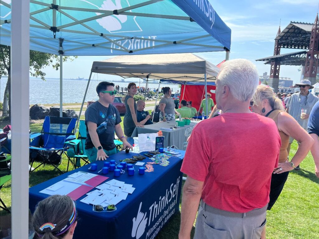 Image of a busy outdoor event scene with people gathered around a booth marked by a banner for ThinkSelf, located in a park by the water. The booth is manned by several individuals in teal shirts, interacting with attendees. This setting appears to be a public outreach or community engagement event.