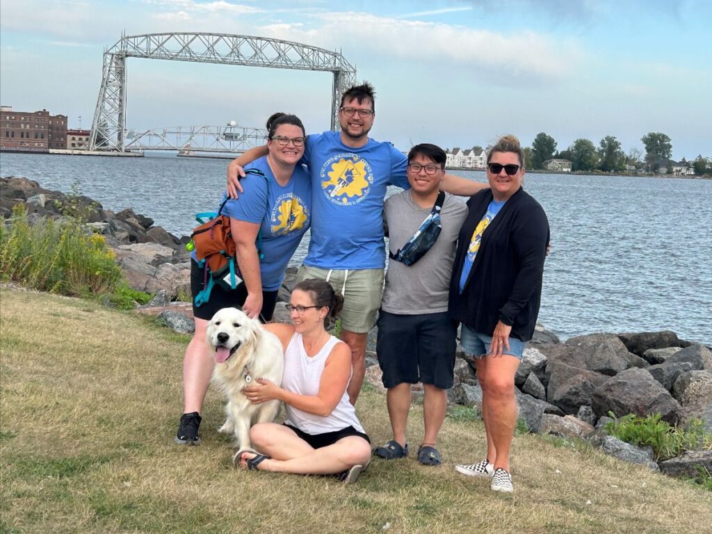 The image features a group of five people and a dog posing together by a lake. They are smiling and seem to be enjoying their time outdoors. In the background, there is a large lift bridge and some buildings across the water.