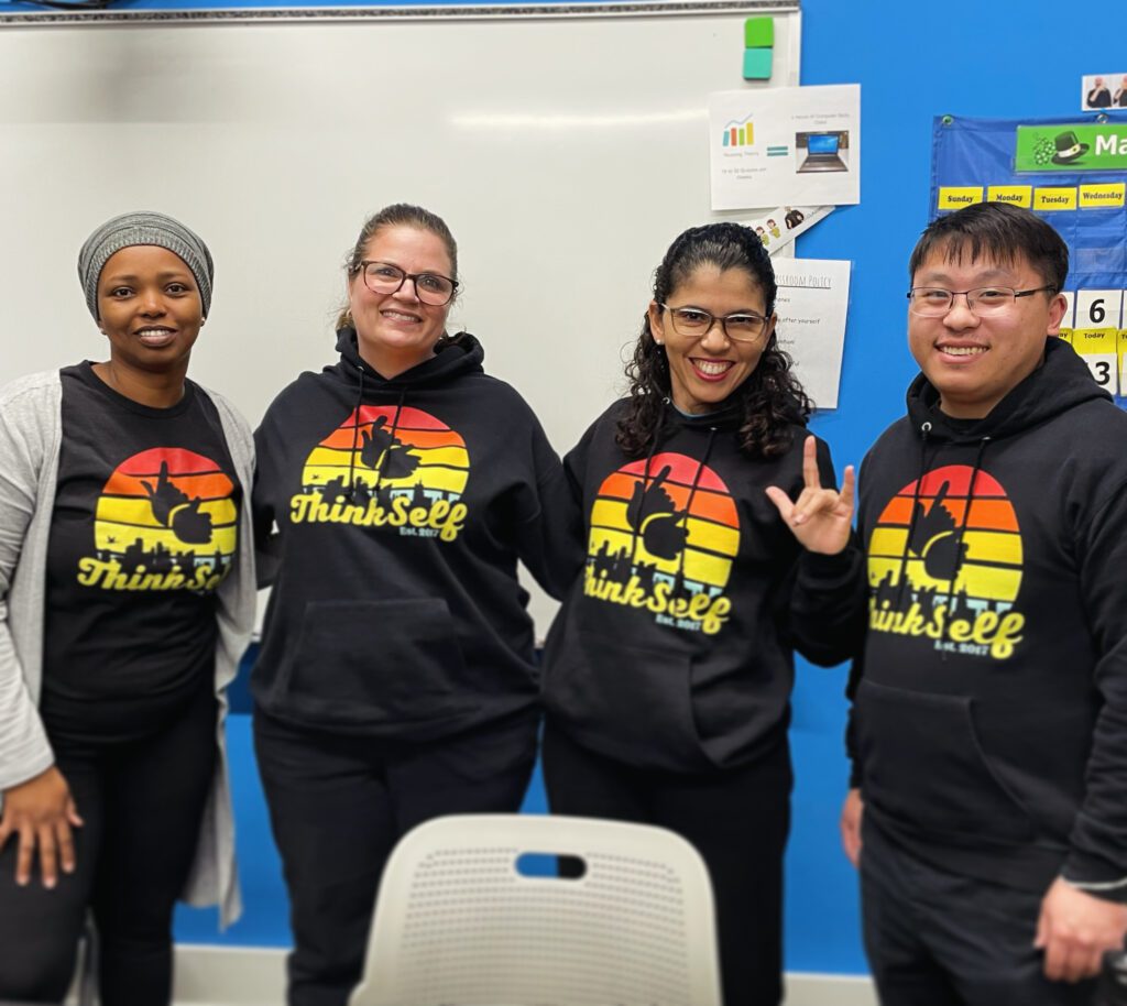 Four people wearing matching black hoodies with the "ThinkSelf" logo in vibrant sunset colors pose in a classroom setting. Each individual is smiling, adding a warm, welcoming vibe to the group photo. The classroom background, complete with educational materials and a whiteboard, indicates that they are likely involved in educational or advocacy efforts related to the ThinkSelf organization. One woman in the group is making a peace sign, further emphasizing the friendly and positive atmosphere of the gathering.