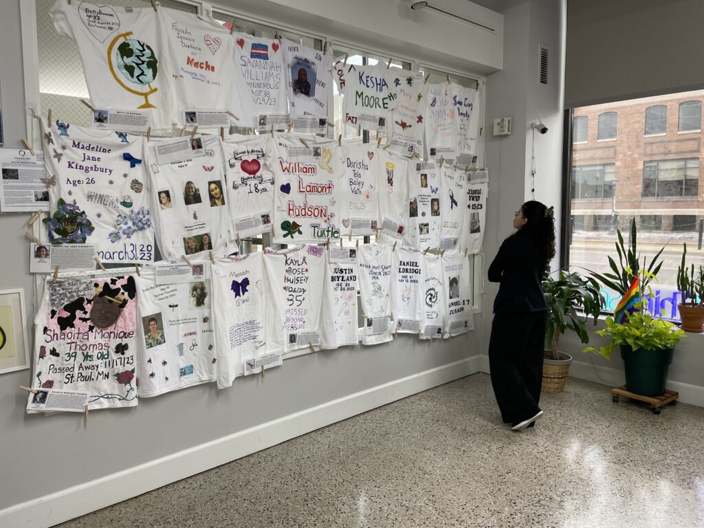 A poignant display of memorial t-shirts pinned to a wall, each decorated with personal messages, photos, and art commemorating lost loved ones. A woman stands to the right, observing the memorials intently, in a reflective posture.