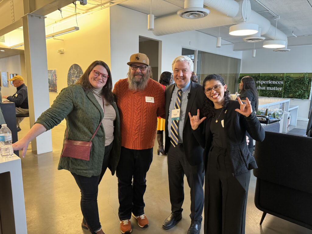 Four people posing happily together at an indoor event, each dressed in smart-casual attire. The modern art gallery setting, featuring a man in the background viewing artworks, adds a cultural backdrop to the gathering. The two women and two men in the foreground smile broadly, one woman making a peace sign with her hands, indicating a lively and joyful interaction. A sign in the background mentions "experiences that matter"