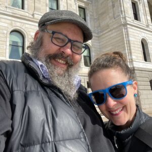 Two people stand close together, smiling. The person on the left has a beard, wears glasses, and a flat cap, while the person on the right wears blue sunglasses. They are outside a light-colored stone building.