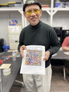 The image shows a man in a black turtleneck and cap, wearing yellow-tinted glasses, holding up a colorful mosaic art piece. He is standing in a room that appears to be a workshop, with shelves and crafting materials in the background.