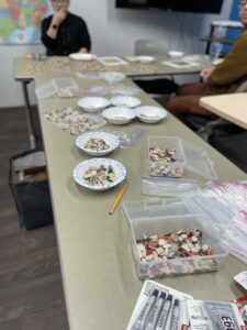 The image displays a classroom or workshop setting with a focus on a table scattered with various crafting materials, including small mosaic tiles spread across multiple plates and containers.