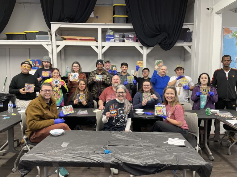 Group photo of participants displaying their mosaic art pieces. They are seated and standing around tables covered with black tablecloths. Most participants are wearing gloves, smiling, and proudly showing their colorful mosaic creations.