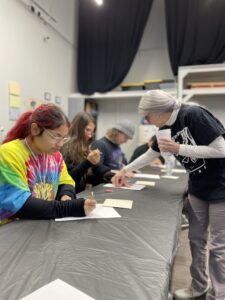 The image captures a group of individuals engaged in an art workshop. They are focused on their tasks, working at a long table covered with a gray tarp. The environment suggests a creative or educational setting, possibly within a studio equipped with supplies and ample lighting for crafting.