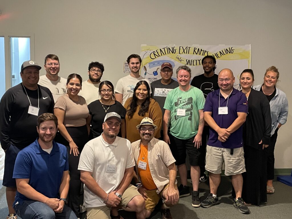 Group photo of diverse individuals, standing and kneeling in two rows. The backdrop includes a poster reading "CREATING EXIT RAMPS INTO SAFETY AND HEALING." All are smiling, some wearing nametags, in casual attire.