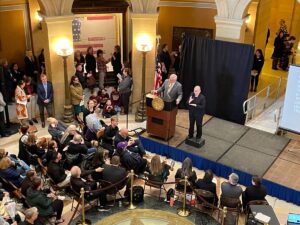 A public event inside a building with ornate columns and lighting. A speaker stands at a podium, with an interpreter beside them. The audience sits and stands around, some are engaged in conversation, and others are watching the speaker.