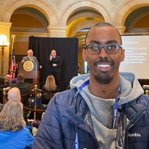 A man with glasses wearing a blue lanyard and jacket stands in the foreground. Behind him, a person is speaking at a podium, accompanied by a sign language interpreter.