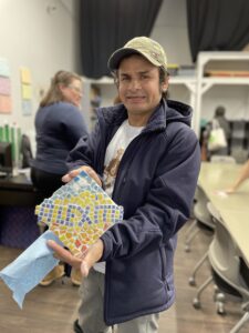 The image shows a man in a blue jacket and a cap, holding a colorful mosaic art piece shaped like a heart. He is in a workshop setting with other people and materials visible in the background.