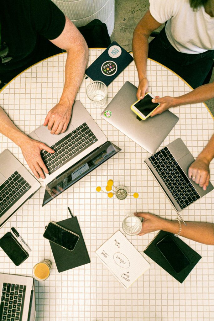 Overhead view of a table with multiple people working on laptops, using phones, and taking notes.